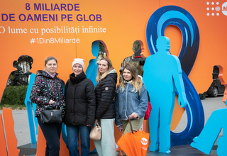 Four refugee women standing in front of the thematic orange stand with key messages in support of the #8BillionStrong campaign