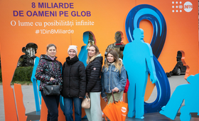 Four refugee women standing in front of the thematic orange stand with key messages in support of the #8BillionStrong campaign